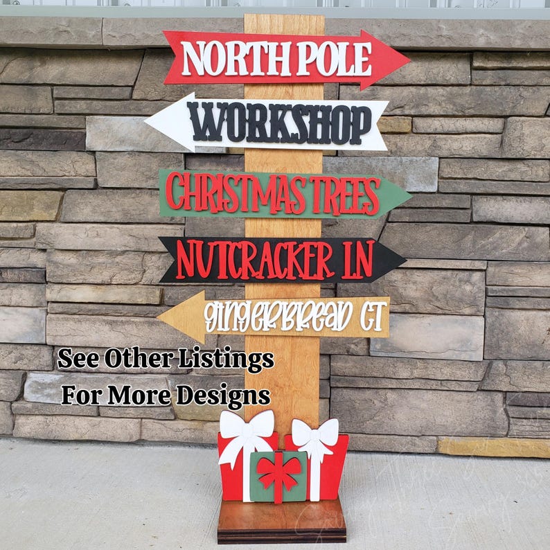 May include: Wooden Christmas-themed directional sign with arrows pointing to "North Pole", "Workshop", "Christmas Trees", "Nutcracker IN", and "Gingerbread CT". The sign includes gift box cutouts at the base and text that reads "See Other Listings For More Designs".
