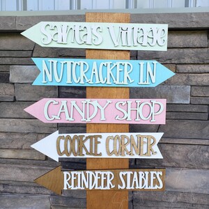 May include: Wooden directional sign with arrows pointing to Santa's Village, Nutcracker Inn, Candy Shop, Cookie Corner, and Reindeer Stables. The arrows are painted in pastel colors with white and brown lettering, mounted on a wooden post.