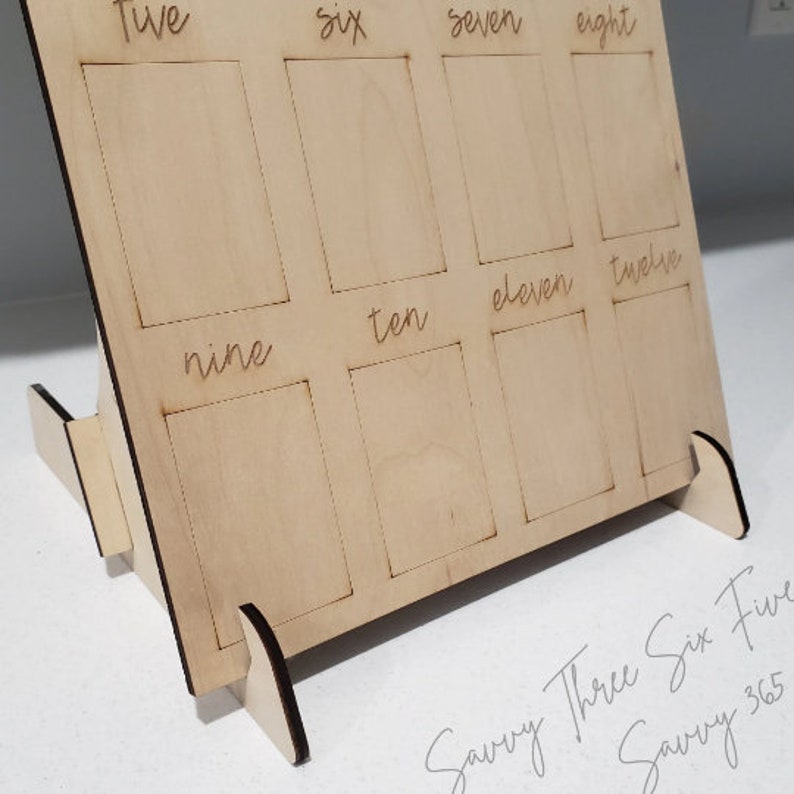 May include: Wooden number learning board with slots for numbers one through twelve. The board is made of light brown wood and has a stand. The numbers are written in cursive.