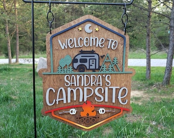 May include: A wooden hanging sign with a dark brown finish featuring the text "WELCOME TO SANDRA'S CAMPSITE". The design includes a layered camper, trees, a crescent moon, stars, and a campfire with two marshmallows on roasting sticks.