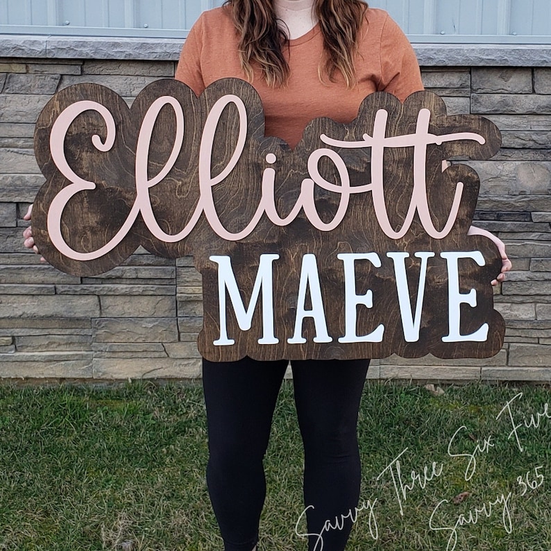 May include: A wooden sign with the names "Elliott" and "Maeve" written in cursive. The name "Elliott" is in pink and the name "Maeve" is in white.