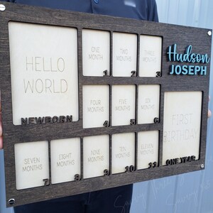 May include: A dark stained wooden milestone board with cutouts for photos. The board includes the text "Hello World, Newborn, First Birthday, One Year" and spaces for monthly photos. The name "Hudson Joseph" is displayed in blue.