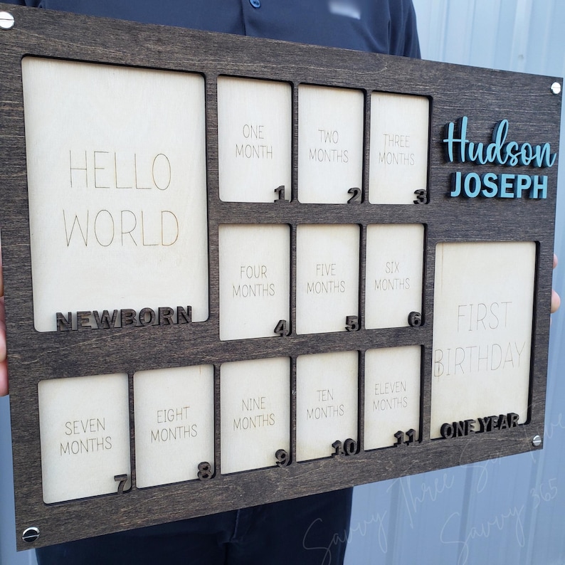 May include: A dark stained wooden milestone board with cutouts for photos. The board includes the text "Hello World, Newborn, First Birthday, One Year" and spaces for monthly photos. The name "Hudson Joseph" is displayed in blue.