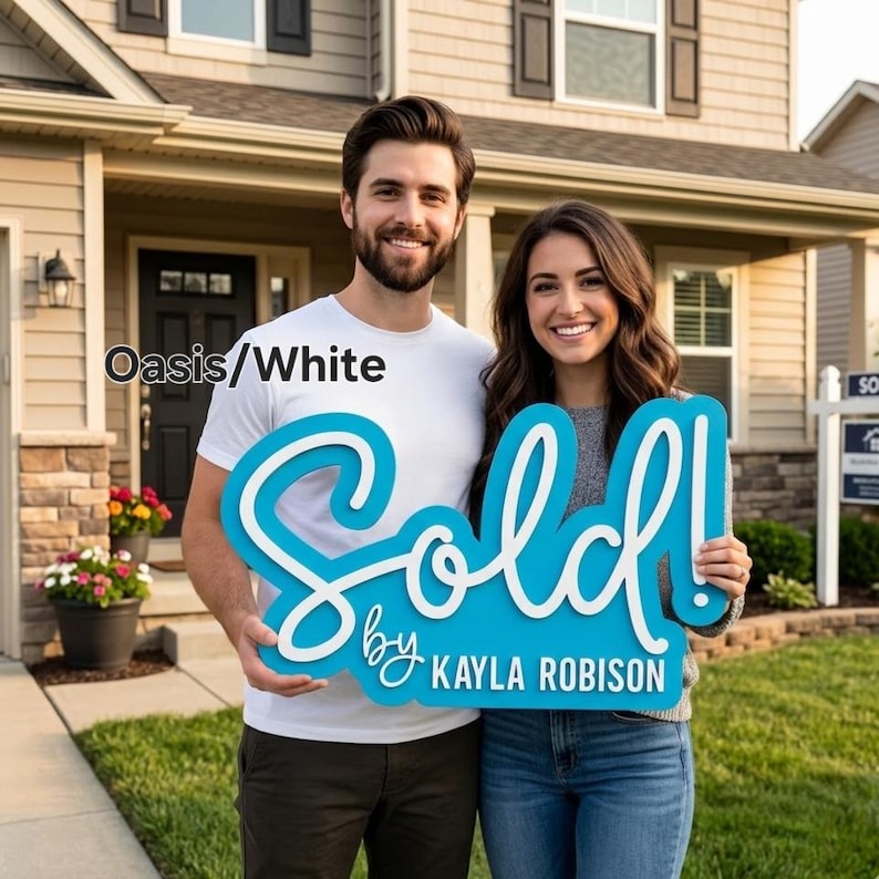 May include: A couple stands in front of a house, holding a large, blue sign that reads "Sold!" in white script. The sign also includes the text "by Kayla Robison." The text "Oasis/White" is in the upper left corner.
