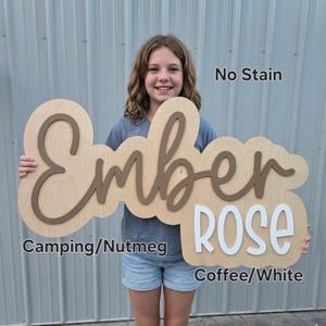 May include: A custom wooden sign featuring the name Ember Rose in layered lettering. The word Ember is in a brown script font, and the word Rose is in a white block font. The sign has a light wood backing with the text No Stain, Camping/Nutmeg, and Coffee/White.