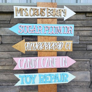 May include: Wooden directional sign with five arrows pointing right. Each arrow is a different color and features a different holiday-themed destination: Mrs. Claus Bakery, Sugar Plum Dr, Gingerbread GT, Candy Cane IN, and Toy Repair.
