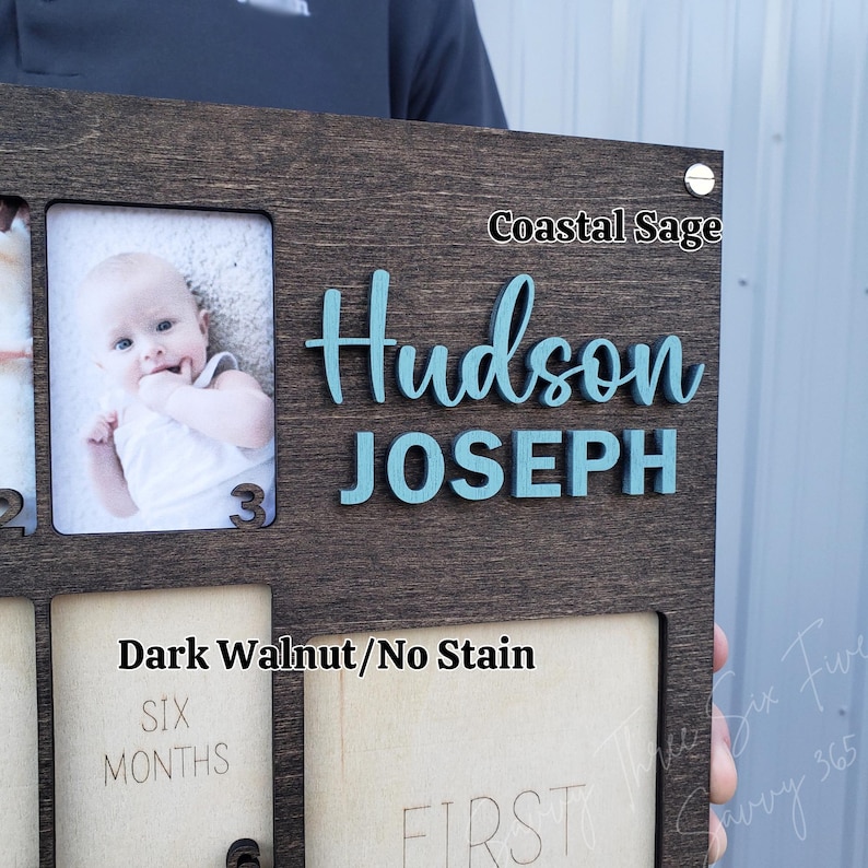May include: A dark walnut wooden photo frame with the name "Hudson JOSEPH" in teal letters. The frame has spaces for photos and the text "SIX MONTHS" and "FIRST". The text "Coastal Sage" is also visible.