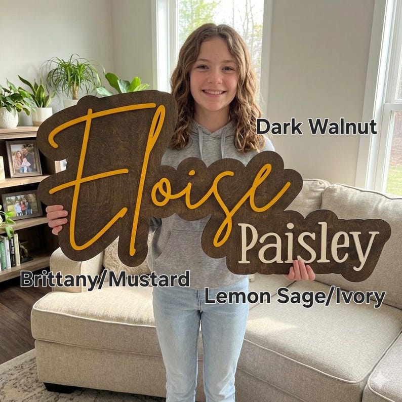 May include: A large wooden wall sign featuring the name Eloise Paisley in layered script and block lettering. The sign has a dark walnut wood backing with mustard yellow script for Eloise and ivory block lettering for the name Paisley.