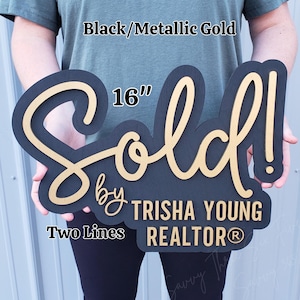 May include: A black and metallic gold "Sold" sign, 16 inches in length, with the text "by Trisha Young Realtor". The sign is two-toned, with a black base and gold lettering. The sign is designed for real estate.