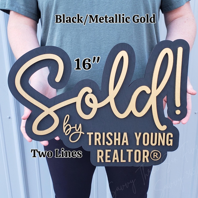 May include: A black and metallic gold "Sold" sign, 16 inches in length, with the text "by Trisha Young Realtor". The sign is two-toned, with a black base and gold lettering. The sign is designed for real estate.