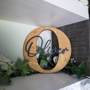 May include: A wooden oval-shaped sign with the name "Oliver" cut out in black lettering. The sign is on a white shelf with green plants and white flowers.