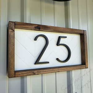 May include: A rectangular wooden address sign with the number 25 in black, mounted on a white chevron patterned background. The frame is a dark brown color. The sign is designed for outdoor use.