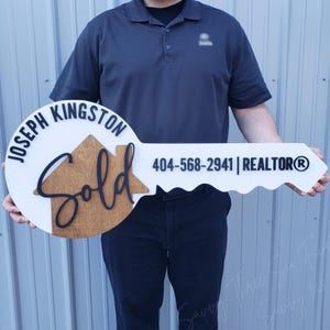 May include: A white and brown wooden key-shaped sign with the text "Joseph Kingston Sold 404-568-2941 | REALTOR®" on it.