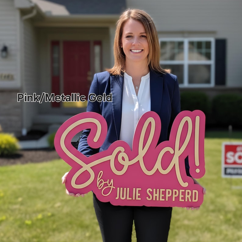 May include: A pink and metallic gold "Sold!" sign with the text "by Julie Shepperd". The sign is held in front of a house. The sign is a real estate marketing tool.