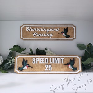 May include: Two wooden signs with white text and a hummingbird graphic. The top sign reads "Hummingbird Crossing". The bottom sign reads "Speed Limit 25".