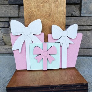 May include: Wooden gift box decorations in pink, white, and light blue. The gifts have white bows and are arranged on a wooden base. The background is a wooden panel and a stone wall.