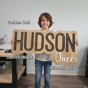 May include: A wooden sign with a golden oak finish featuring the name HUDSON in large black block letters and the name Jace in white script lettering. The sign has a scalloped edge design and includes text labels for Golden Oak, Ostrich/Black, and Rihana/White.