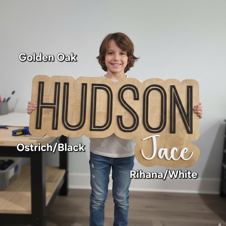 May include: A wooden sign with a golden oak finish featuring the name HUDSON in large black block letters and the name Jace in white script lettering. The sign has a scalloped edge design and includes text labels for Golden Oak, Ostrich/Black, and Rihana/White.