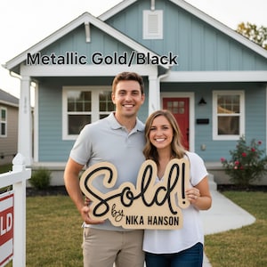May include: A couple stands in front of a blue house, holding a wooden sign that reads "Sold!" in black script. The sign also includes "by Nika Hanson." The text "Metallic Gold/Black" is at the top of the image.