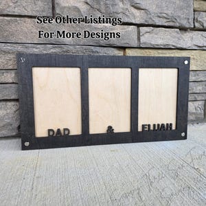 May include: A black wooden picture frame with three openings. The frame has the words "DAD" and "ELIJAH" cut out, with an ampersand symbol between them. The text "See Other Listings For More Designs" is at the top.
