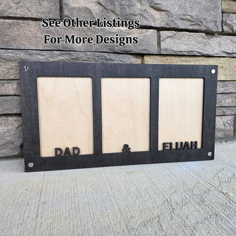 May include: A black wooden picture frame with three openings. The frame has the words "DAD" and "ELIJAH" cut out, with an ampersand symbol between them. The text "See Other Listings For More Designs" is at the top.