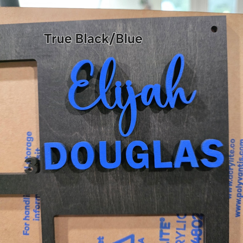 May include: A black wooden sign with the name "Elijah" in blue cursive letters above the name "DOUGLAS" in blue block letters. The text "True Black/Blue" is at the top.