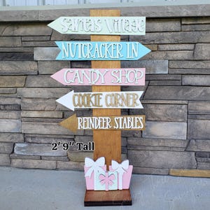 May include: Wooden directional sign with arrows pointing to Santa's Village, Nutcracker Inn, Candy Shop, Cookie Corner, and Reindeer Stables. The sign is 2 feet 9 inches tall and features a base with gift box cutouts. The sign is made of wood and painted in pastel colors.