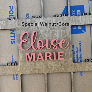 May include: A wooden frame with cut-out sections, featuring the name "Eloise Marie" in pink script and block letters. The frame is labeled "Special Walnut/Coral". Numbers and text are visible in the background.