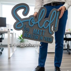 May include: A carbon gray sign with the word "Sold!" in a script font, accented with lagoon blue. The sign also includes the text "by MATT REALTOR IN TECH".