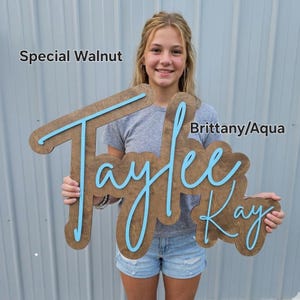 May include: A custom wooden name sign featuring the name Taylee Kay in a script font. The base is a dark brown Special Walnut wood finish, and the raised lettering is in a bright aqua colour. The text Special Walnut and Brittany/Aqua is visible.
