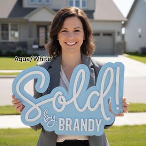 May include: An aqua and white "Sold" sign with the text "by BRANDY". The sign is held in front of a house. The sign is a decorative item for real estate.