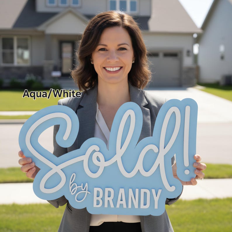 May include: An aqua and white "Sold" sign with the text "by BRANDY". The sign is held in front of a house. The sign is a decorative item for real estate.