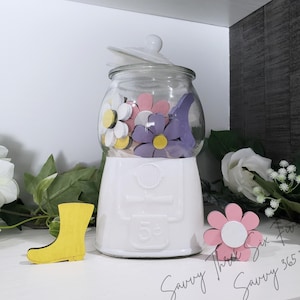 May include: White ceramic gumball machine with a glass jar filled with colorful wooden flowers. The machine has a 5 cent sign on the front. There is a yellow wooden boot and a pink wooden flower on the table.