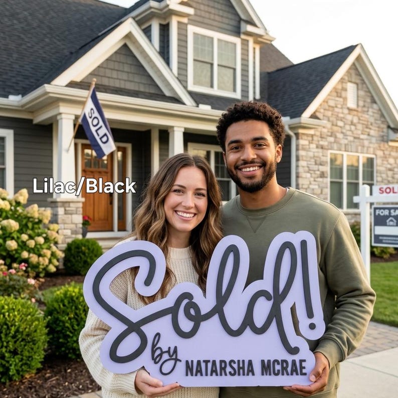 May include: A couple smiles in front of a house, holding a sign that says "Sold!" in a cursive font. The sign is lilac with black lettering and the realtor's name. A "Sold" flag flies in the background.