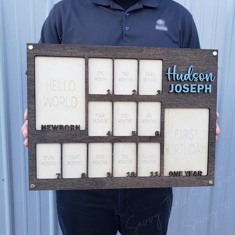 May include: A dark wooden baby milestone board with spaces for photos and text. The board includes the text "Hello World," "Newborn," and "First Birthday." It also features the name "Hudson Joseph" in blue.
