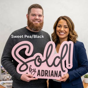 May include: A pink and black sign that says "Sold! by Adriana" is held by two people. The sign is in a cursive font with a black outline and the background is pink. The text "Sweet Pea/Black" is in the upper left corner.