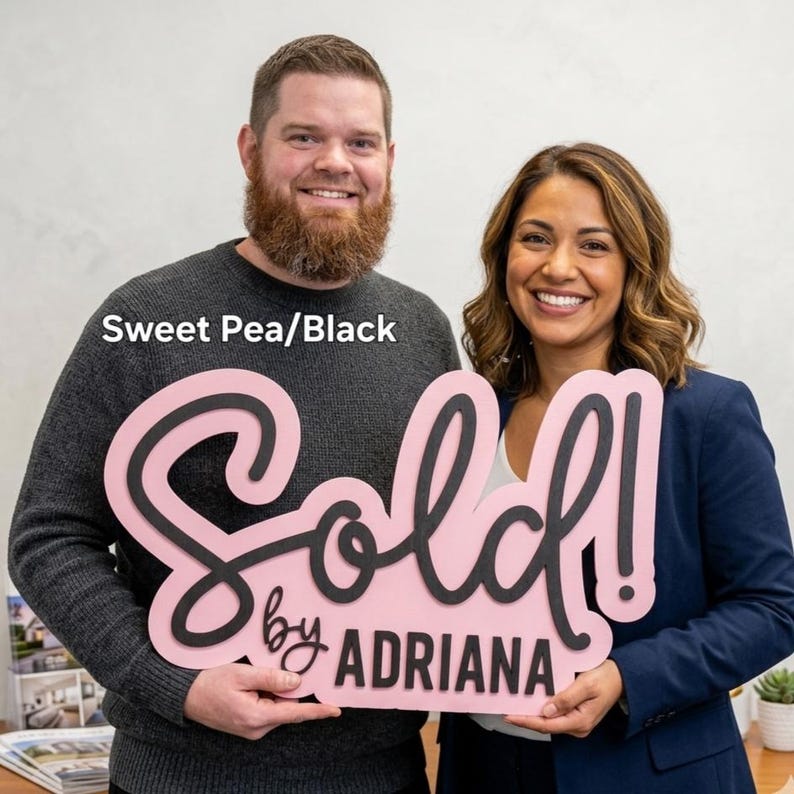 May include: A pink and black sign that says "Sold! by Adriana" is held by two people. The sign is in a cursive font with a black outline and the background is pink. The text "Sweet Pea/Black" is in the upper left corner.