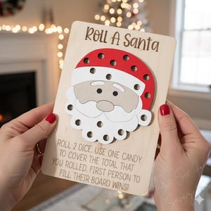 May include: A wooden Christmas game board featuring a Santa Claus design with a red hat and white beard. The text "Roll A Santa" is at the top, with instructions below. The game board is held by a person with red nail polish.
