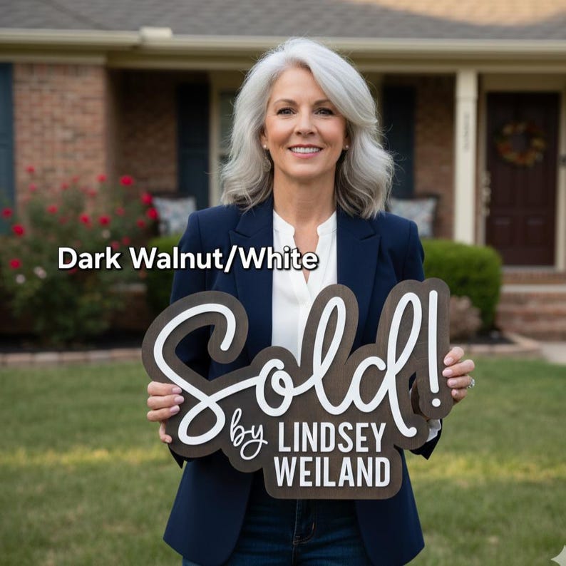 May include: A dark walnut and white wooden sign that says "Sold!" with the text "by Lindsey Weiland". The sign is held in front of a house by a person wearing a navy blue blazer.