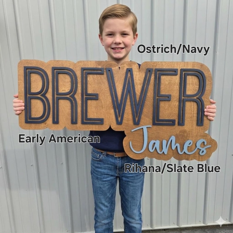 May include: A custom wooden sign featuring the name BREWER in navy blue block letters on a brown wood background, with the name James in slate blue script below. Text labels include Ostrich/Navy, Early American, and Rihana/Slate Blue.