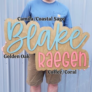 May include: A wooden sign with a golden oak finish featuring the name Blake in light blue script and the name RAEGEN in coral block letters. Text overlays include Camila/Coastal Sage, Golden Oak, and Coffee/Coral identifying the colours.