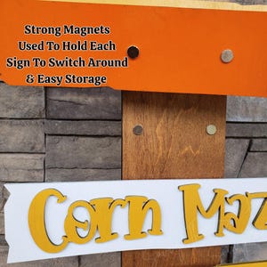 May include: Wooden sign with the words "Corn Maze" in yellow letters on a white background. The sign is attached to an orange and brown wooden frame. The text "Strong Magnets Used To Hold Each Sign To Switch Around & Easy Storage" is also visible.