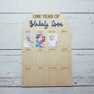 May include: A wooden photo display board with a rounded arch top. The board is designed to hold photos of a baby's first year, with spaces for 12 photos. The board is labeled "One Year of Blakely Rose" and has the numbers one through twelve printed below each photo space.