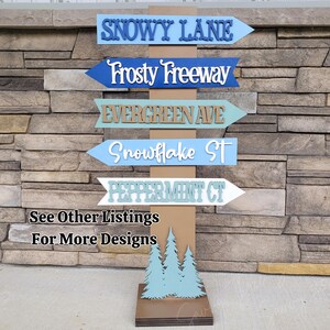 May include: A wooden signpost with five directional signs, each pointing in a different direction. The signs are painted in various colors, including blue, green, and white, and feature names like "Snowy Lane" and "Frosty Freeway." The base of the signpost has a tree design and the text "See Other Listings For More Designs."