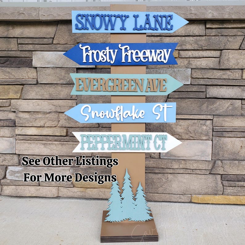 May include: A wooden signpost with five directional signs, each pointing in a different direction. The signs are painted in various colors, including blue, green, and white, and feature names like "Snowy Lane" and "Frosty Freeway." The base of the signpost has a tree design and the text "See Other Listings For More Designs."