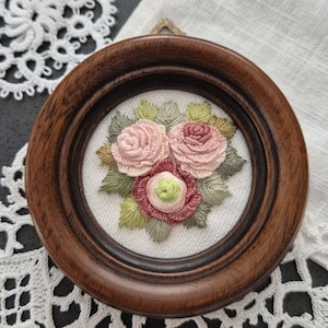Könnte beinhalten: Kleine, runde, gerahmte Stickerei mit einem Strauß Rosen in Rosa- und Rottönen, umgeben von grünen Blättern. Der Rahmen ist aus dunklem Holz. Die Stickerei befindet sich auf weißem Hintergrund.