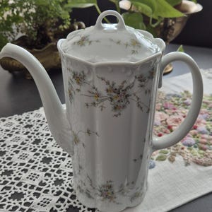 Könnte beinhalten: Weiße Porzellan-Kaffeekanne mit floralem Muster. Die Kanne hat einen langen, gebogenen Ausguss, einen Henkel und einen Deckel. Das Blumenmuster umfasst gelbe, rosa und grüne Blumen. Sie steht auf einem Tisch mit einer weißen Spitzendecke und einem floral bestickten Tischläufer.