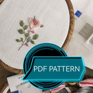 Könnte beinhalten: Nahaufnahme eines Stickrahmens mit floralem Design. Das Design zeigt eine rosa Rose mit grünen Blättern und grauen Akzenten. Der Text "PDF PATTERN" wird in einer blau-schwarzen Grafik angezeigt. Zusätzliches Stickzubehör ist sichtbar.