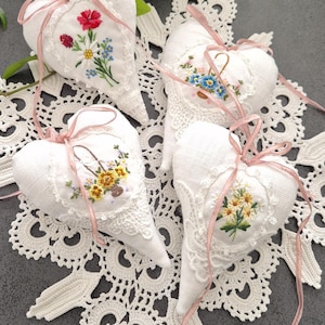 Peut inclure: Quatre sachets blancs en forme de cœur avec des motifs floraux brodés, bordés de dentelle et noués avec des rubans roses. Les sachets sont disposés sur un napperon en dentelle blanche, avec un feuillage vert en arrière-plan.