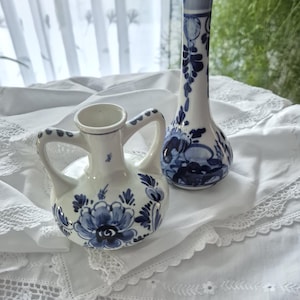 Könnte beinhalten: Zwei weiße Keramikvasen mit blauen Blumenmustern. Eine Vase ist ein kleiner Krug mit Henkeln, die andere eine hohe, schlanke Vase. Beide sind mit blauen Blumenmustern verziert. Sie stehen auf einem weißen, spitzenbesetzten Tuch.